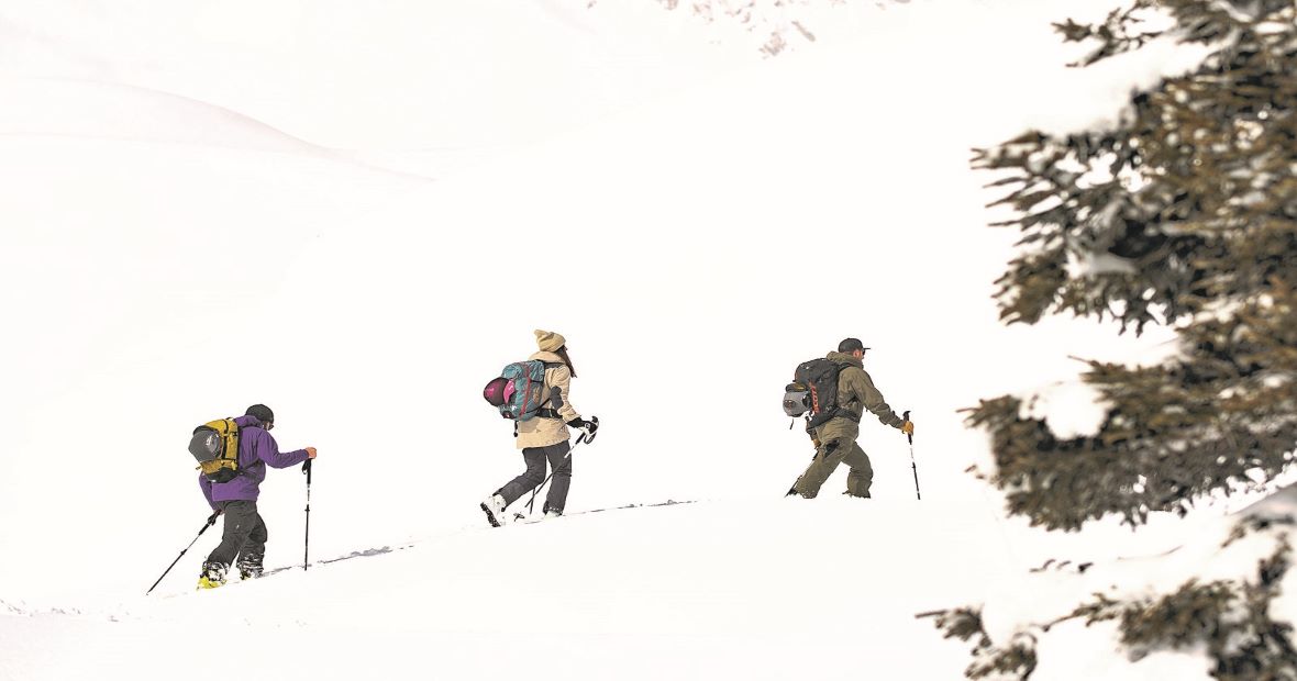 Drei Personen mit Ski- und Bergsteigerausrüstung wandern in einer Schneelandschaft. Die Szene zeigt sie in einer Reihe, während sie mit Stöcken durch den tiefen Schnee gehen. Rechts im Bild ist ein schneebedeckter Baum zu sehen.